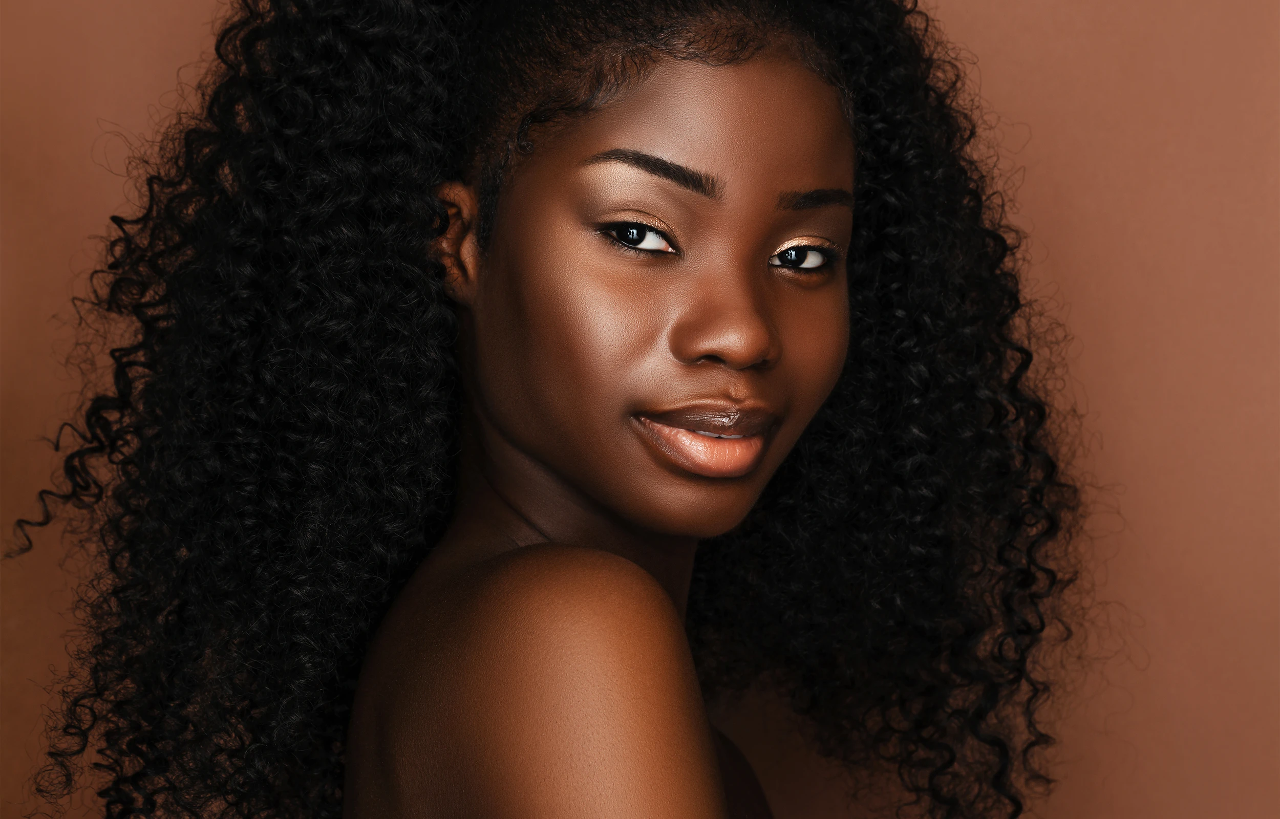 Close-up portrait of a woman with glowing skin and natural curls against a warm brown background